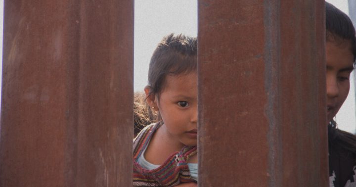 A little girl looks through slats in the US-Mexico border wall.