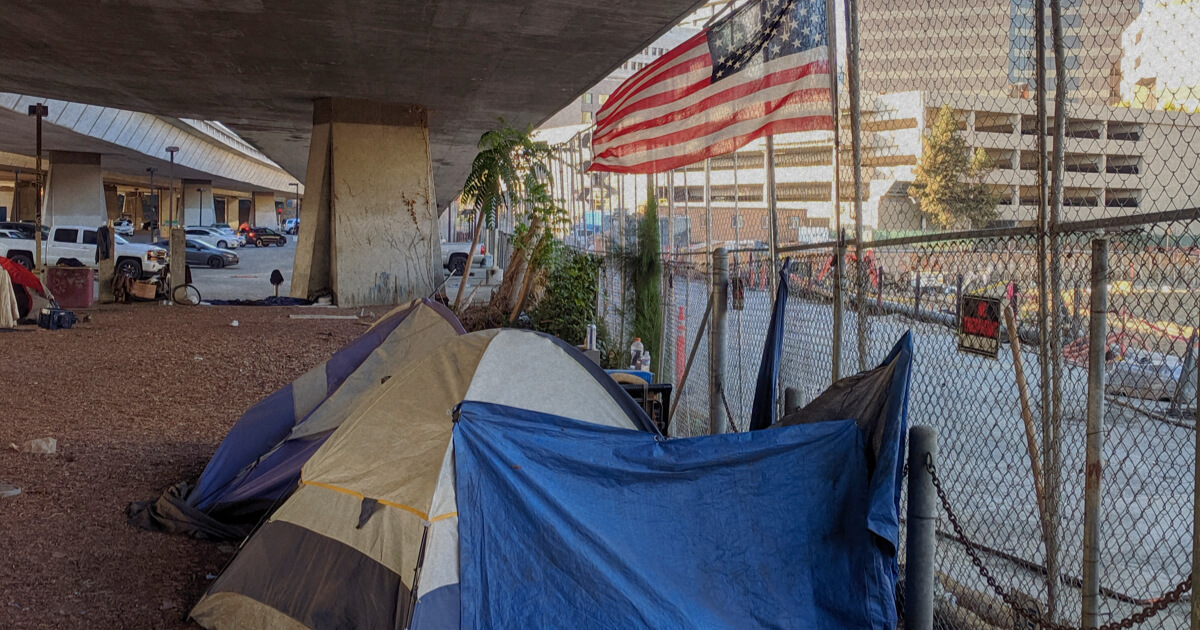 Homeless camp under a bridge with a US flag in the background.